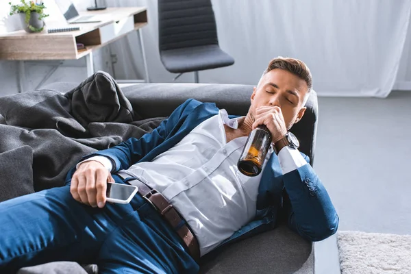 Подавленный бизнесмен пьет алкоголь из бутылки и лежит на диване в гостиной — стоковое фото