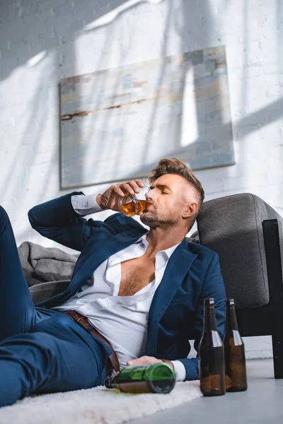 Empresario deprimido beber whisky cerca de botellas en la sala de estar - foto de stock