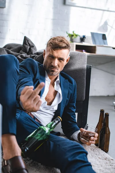 Hombre de negocios borracho mostrando el dedo medio y sosteniendo vidrio con bebida alcohólica - foto de stock