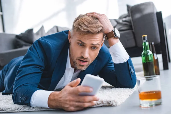 Шокований бізнесмен дивиться на смартфон і лежить на килимі біля скла з алкогольним напоєм — стокове фото
