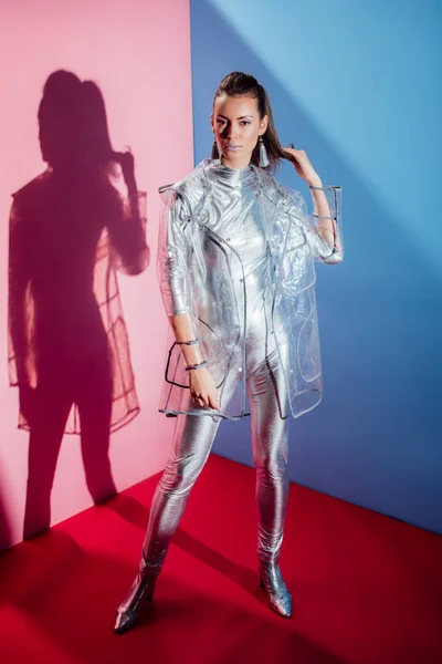 Attractive stylish woman posing in silver bodysuit and raincoat on pink and blue background — Stock Photo