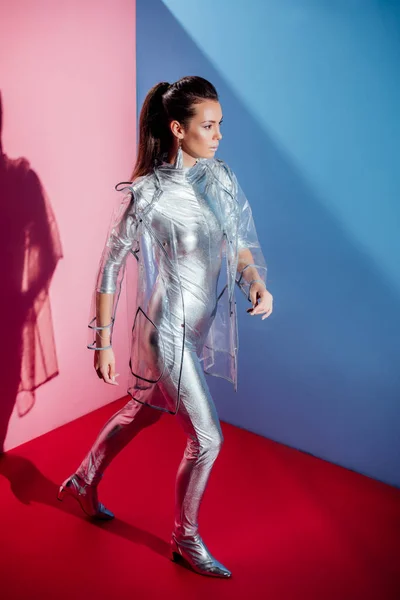 Hermoso modelo elegante posando en traje de cuerpo de plata y impermeable sobre fondo rosa y azul - foto de stock