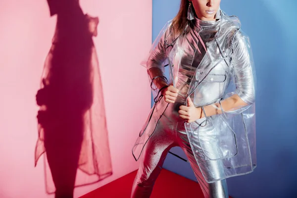 Cropped view of stylish woman posing in silver bodysuit and raincoat on pink and blue background — Stock Photo