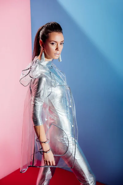 Fashionable young woman posing in silver bodysuit and raincoat on pink and blue background — Stock Photo
