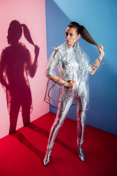 Stylish girl posing in metallic bodysuit and trendy raincoat for fashion shoot on pink and blue background — Stock Photo