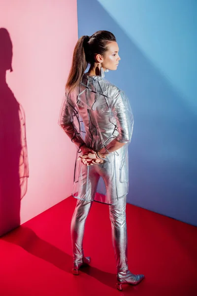 Back view of fashionable woman posing in silver bodysuit and raincoat on pink and blue background — Stock Photo