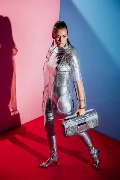 Attractive woman in metallic bodysuit and raincoat posing with boombox on pink and blue background — Stock Photo