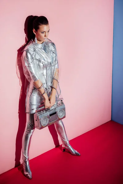 Elegant model in metallic bodysuit and raincoat posing with boombox on pink and blue background — Stock Photo