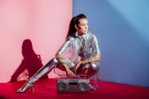 Fashionable model in silver bodysuit and raincoat posing with boombox on pink and blue background — Stock Photo