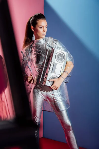 Mujer joven de moda en traje de cuerpo metálico y impermeable posando con boombox retro sobre fondo rosa y azul - foto de stock