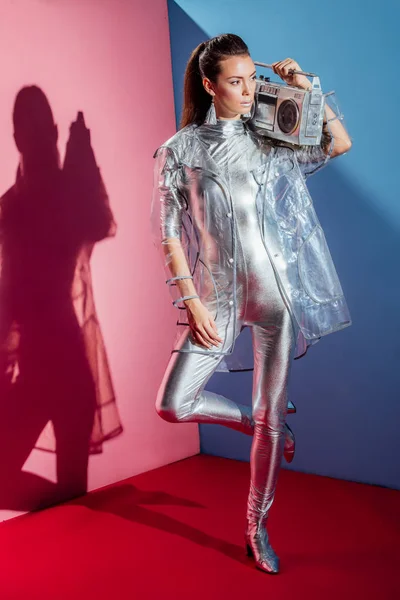 Attraente ragazza elegante in tuta metallica e impermeabile in posa con retro boombox su sfondo rosa e blu — Foto stock