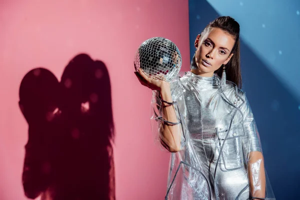 Modelo de moda en traje de cuerpo de plata y impermeable posando con bola disco sobre fondo rosa y azul - foto de stock