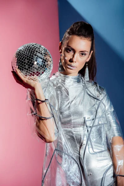 Modelo bonito em bodysuit prata e capa de chuva posando com bola de discoteca no fundo rosa e azul — Fotografia de Stock