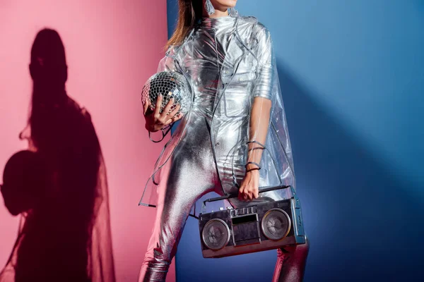 Vue recadrée de la femme à la mode en costume de corps argenté et imperméable posant avec boombox et boule disco sur fond rose et bleu — Photo de stock