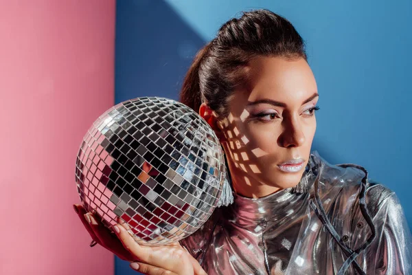 Beautiful stylish young woman posing with silver disco ball on pink and blue background — Stock Photo