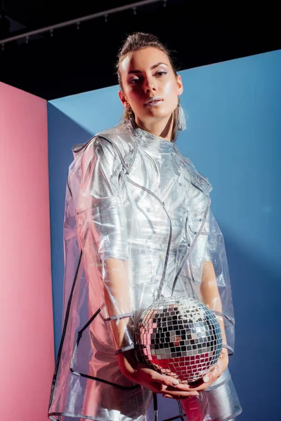 Beautiful elegant girl in silver bodysuit and raincoat posing with disco ball on pink and blue background — Stock Photo