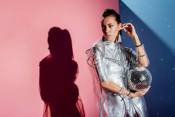 Mujer joven con estilo en traje de cuerpo metálico y impermeable posando con bola disco sobre fondo rosa y azul - foto de stock