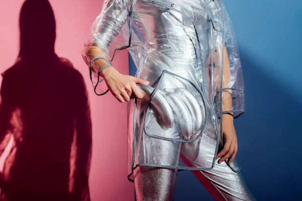 Partial view of elegant girl in metallic bodysuit and fashionable raincoat posing with silver bananas for fashion shoot on pink and blue background — Stock Photo