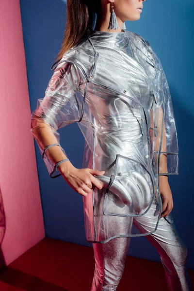 Vista recortada de chica en traje de cuerpo metálico y impermeable posando con plátanos plateados para disparar de moda sobre fondo rosa y azul - foto de stock
