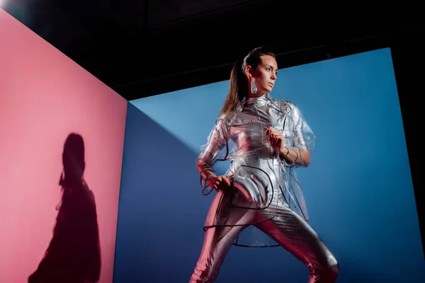 Beautiful fashionable woman in metallic bodysuit and raincoat posing with silver bananas on pink and blue background — Stock Photo