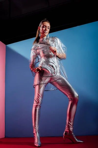 Mujer joven de moda en traje de cuerpo metálico y impermeable posando con plátanos plateados sobre fondo azul - foto de stock