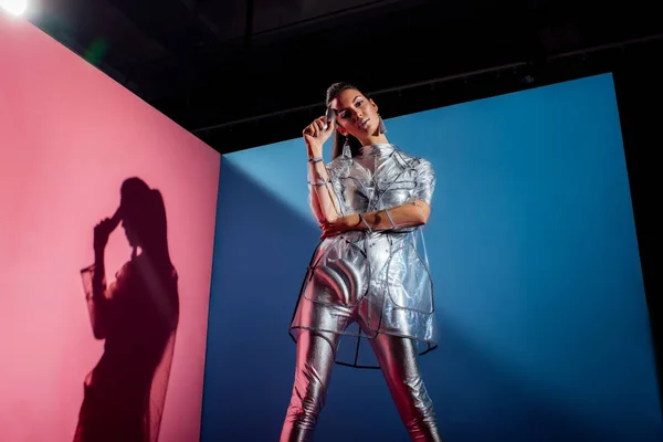 Beautiful stylish young woman in metallic bodysuit and raincoat posing with silver bananas on pink and blue background — Stock Photo