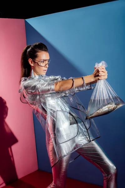 Trendy girl in silver bodysuit and raincoat holding plastic bag with fish on pink and blue background — Stock Photo
