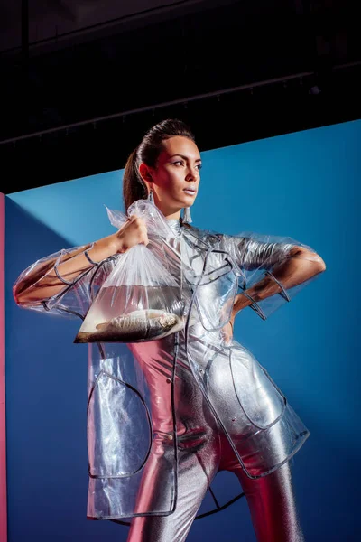 Hermoso modelo en traje de cuerpo de plata y impermeable con bolsa de plástico con peces sobre fondo azul - foto de stock