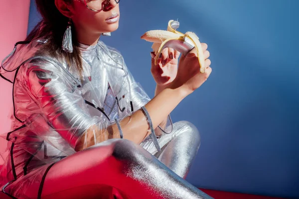 Vista recortada de la mujer joven de moda en traje de cuerpo metálico y impermeable posando con plátano sobre fondo azul - foto de stock