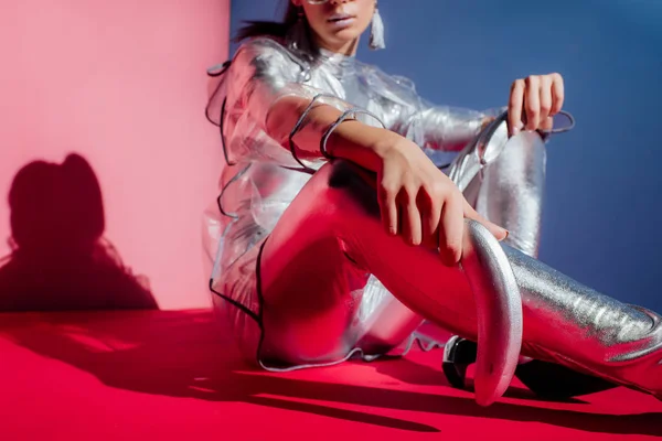 Vista cortada de mulher nova na moda em bodysuit metálico e capa de chuva posando com banana de prata no fundo rosa e azul — Fotografia de Stock