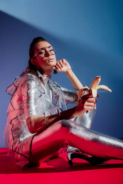 Attractive fashionable girl in metallic bodysuit and raincoat posing with silver banana on pink and blue background — Stock Photo