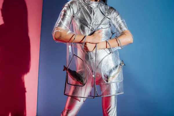 Обрезанный вид на модную модель в серебряном боди и дождевике, позирующую с рыбой в карманах на розовом и синем фоне — стоковое фото