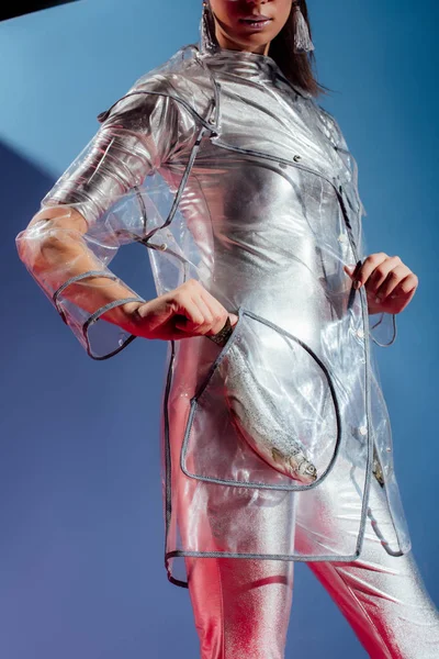 Partial view of fashionable model in silver bodysuit and raincoat posing with fish in pocket on blue background — Stock Photo