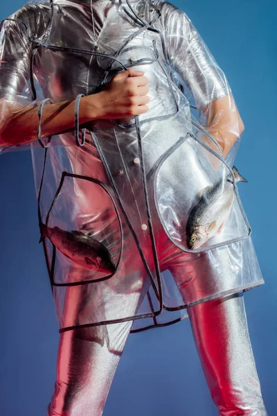 Cropped view of fashionable girl in silver bodysuit posing with fish in pockets of raincoat on blue background — Stock Photo