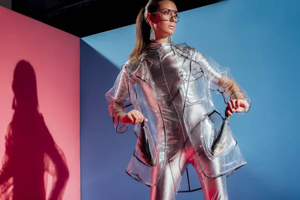 Beautiful stylish girl in silver bodysuit and raincoat posing with fish on pink and blue background — Stock Photo