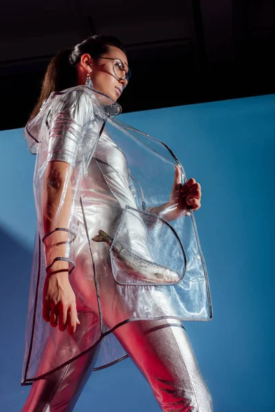 Vista inferior de chica atractiva en traje de cuerpo de plata y impermeable posando con peces sobre fondo azul - foto de stock