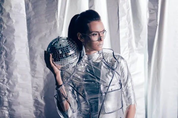 Jeune femme à la mode en costume de corps argenté et imperméable posant avec boule disco sur fond métallique — Photo de stock