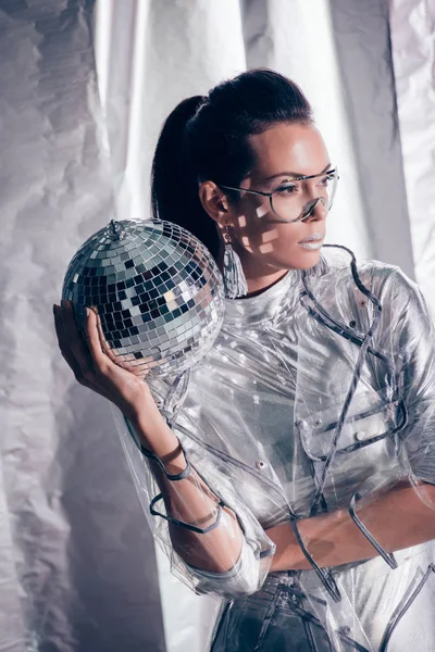 Beautiful stylish young woman in silver bodysuit and raincoat posing with disco ball on metallic background — Stock Photo