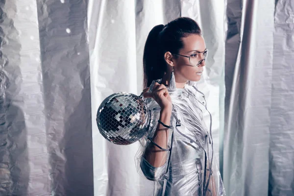 Atractiva modelo joven en traje de cuerpo de plata y impermeable posando con bola disco sobre fondo metálico - foto de stock