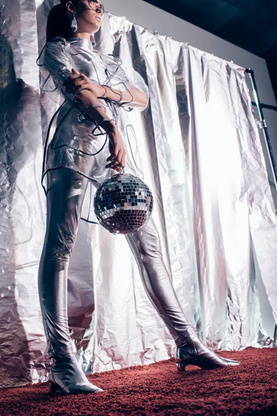 Bottom view of fashionable girl in silver bodysuit and raincoat posing with disco ball on metallic background — Stock Photo