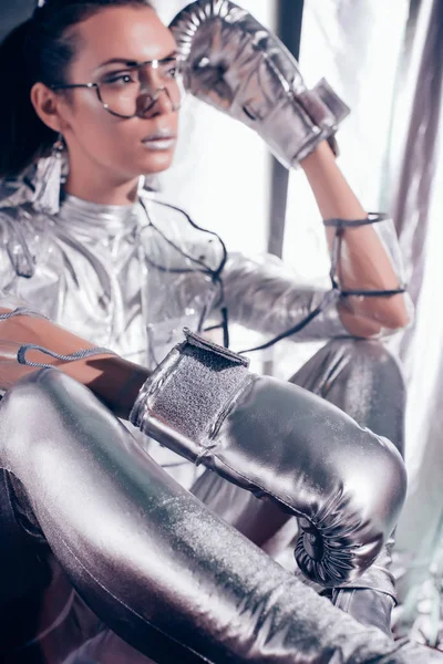 Selective focus of attractive model posing in bodysuit, raincoat and silver box gloves on metallic background — Stock Photo