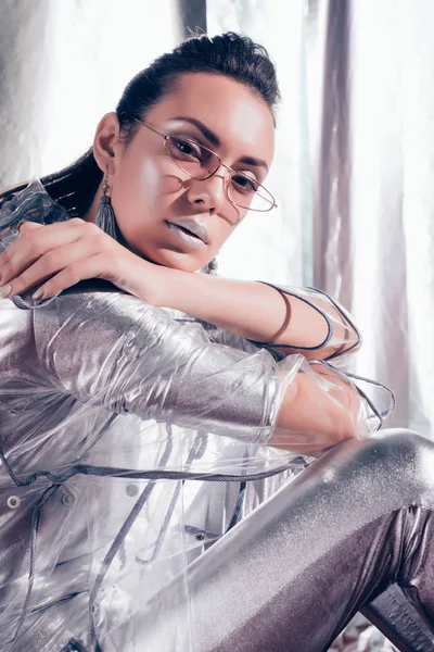 Fashionable girl posing in trendy glasses, silver bodysuit and raincoat on metallic background — Stock Photo