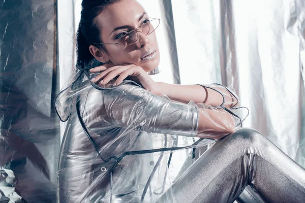 Attraktive junge Frau posiert mit trendiger Brille, silbernem Body und Regenmantel auf metallischem Hintergrund — Stockfoto