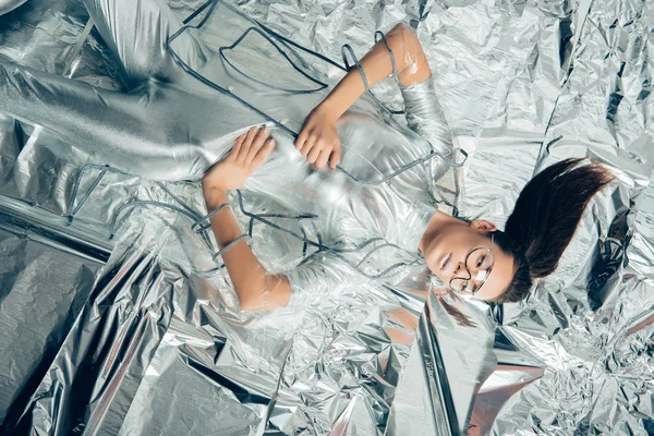 Top view of attractive fashionable girl posing in silver bodysuit and raincoat on metallic background — Stock Photo