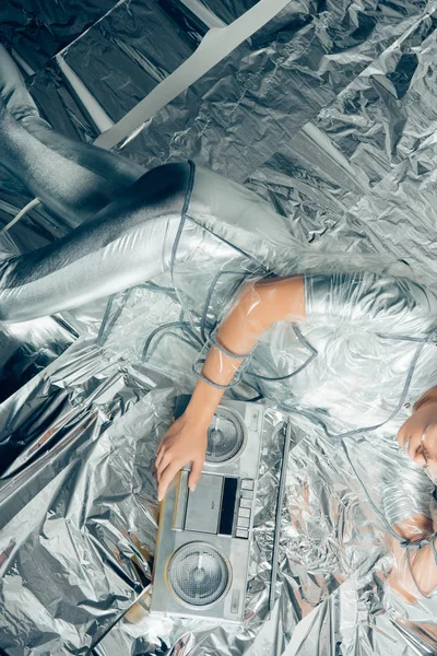 Cropped top view of stylish girl in silver bodysuit and raincoat posing with boombox on metallic background — Stock Photo