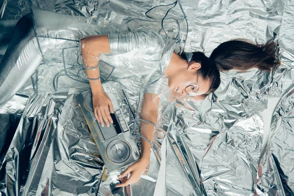 Top view of elegant woman in silver bodysuit and raincoat posing with retro boombox on metallic background — Stock Photo