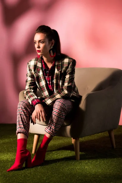 Beautiful young woman in checkered suit posing in armchair on pink background — Stock Photo