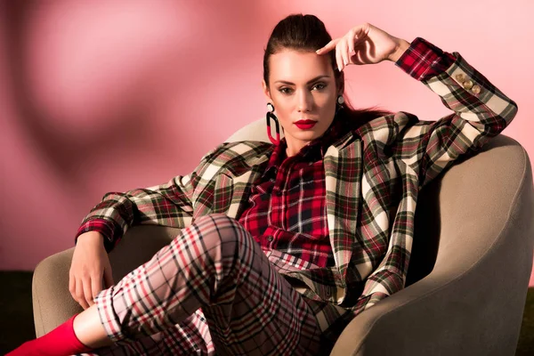 Mujer de moda en traje a cuadros posando en sillón sobre fondo rosa - foto de stock