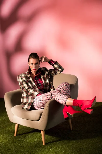 Красива елегантна дівчина в картатому костюмі позує в кріслі на рожевому фоні — стокове фото
