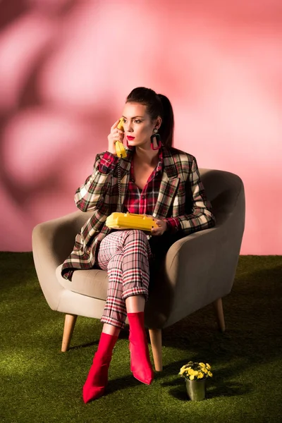 Модная девушка в клетчатом костюме позирует в кресле с ротационным телефоном на розовом фоне — стоковое фото